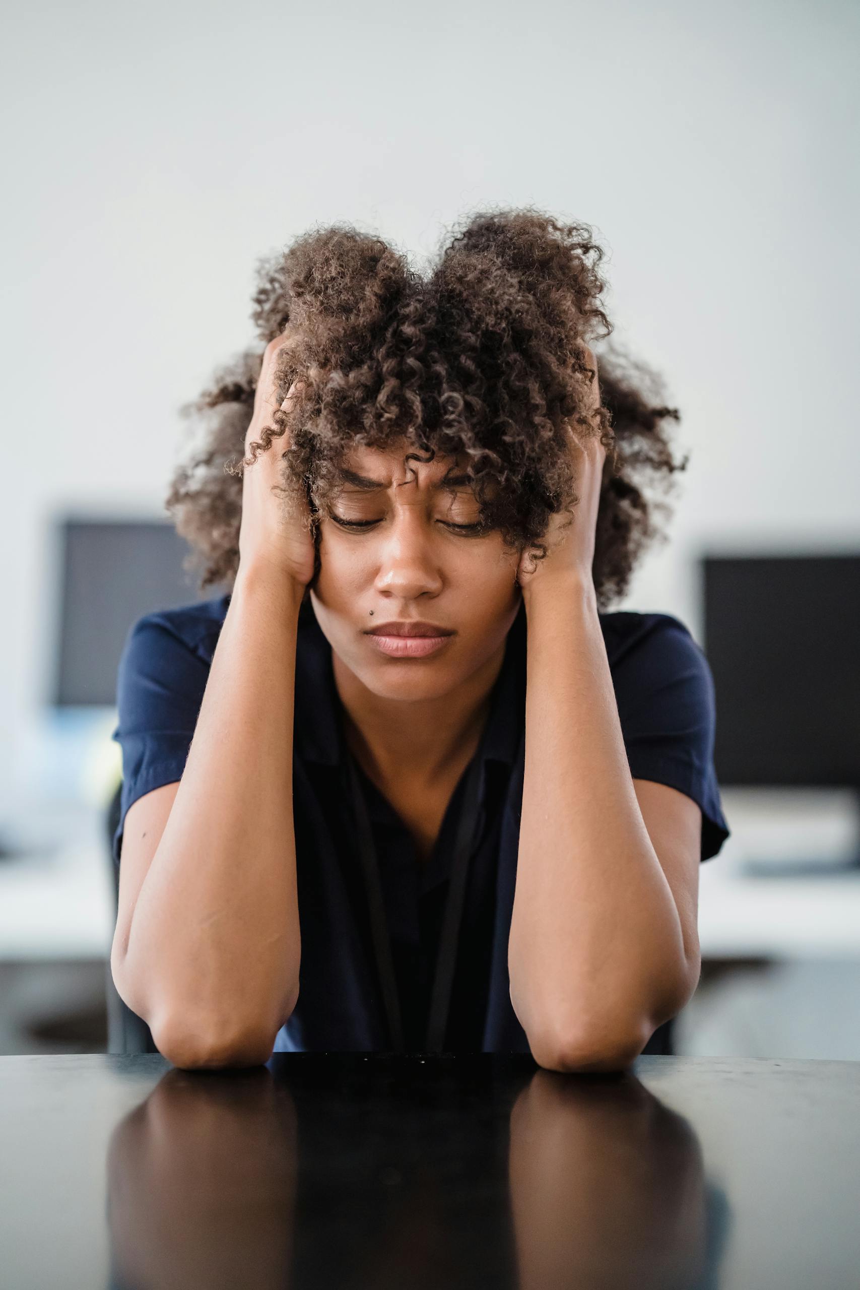 Woman in office setting expresses stress, seated at desk with hands in hair. Perfect for workplace stress concept.